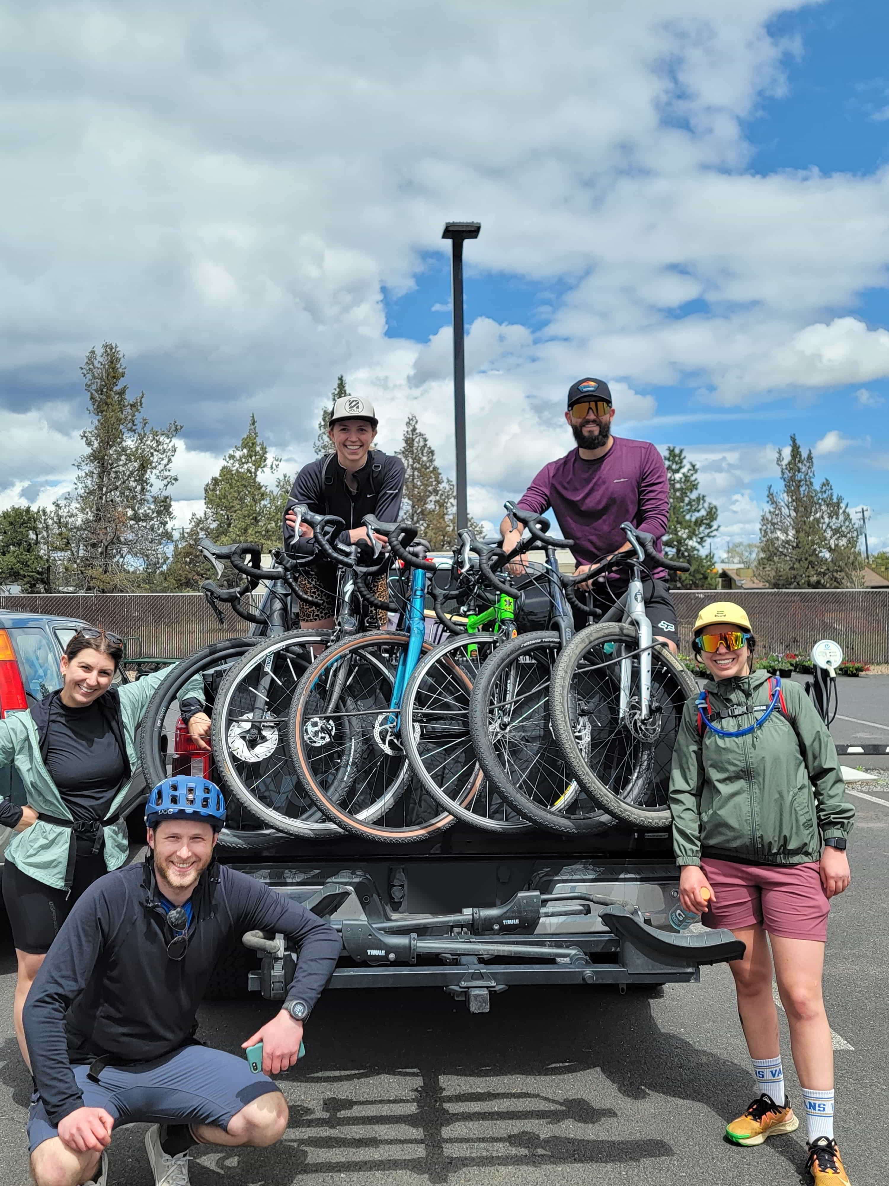 5 people pose on the back of a truck, with bikes in the back. They are all smiling and appear to be having a great time.